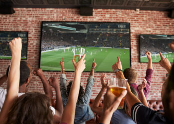 Rear View Of Friends Watching Game In Sports Bar On Screens